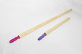 Two long light wood poles with colored plastic ends, one pink and one purple, laid diagonally on a white fabric background.